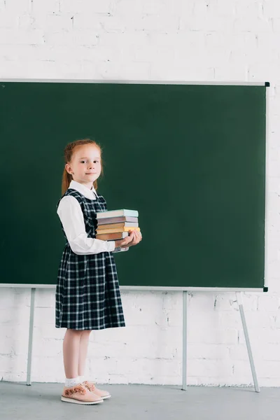 Adorable Little Uczennica Gospodarstwa Stos Książek Patrząc Kamery Stojąc Pobliżu — Zdjęcie stockowe