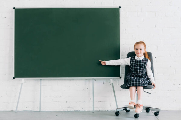 adorable little schoolgirl smiling at camera and pointing at blank chalkboard while sitting on chair
