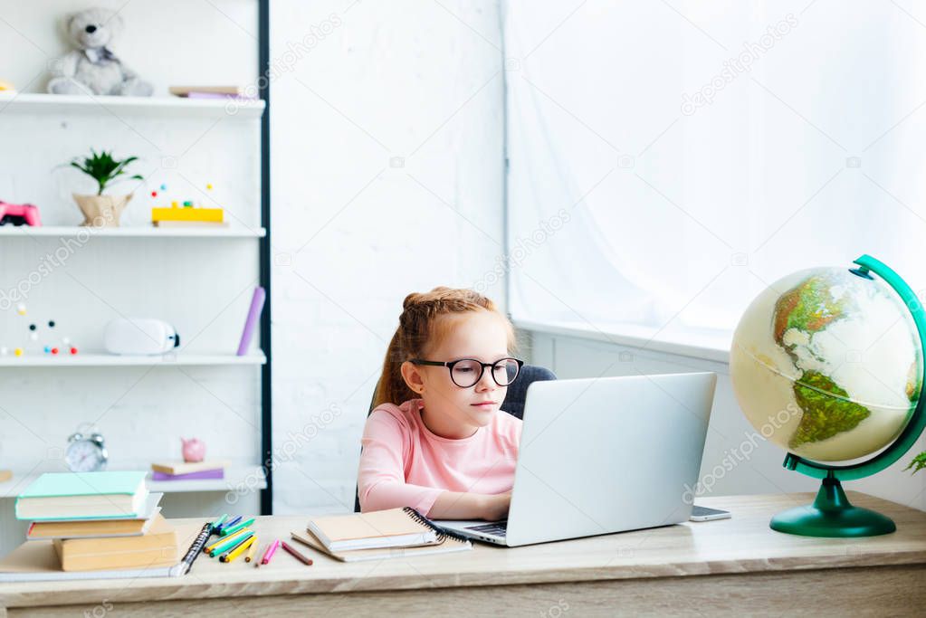 beautiful child in eyeglasses using laptop while studying at desk at home