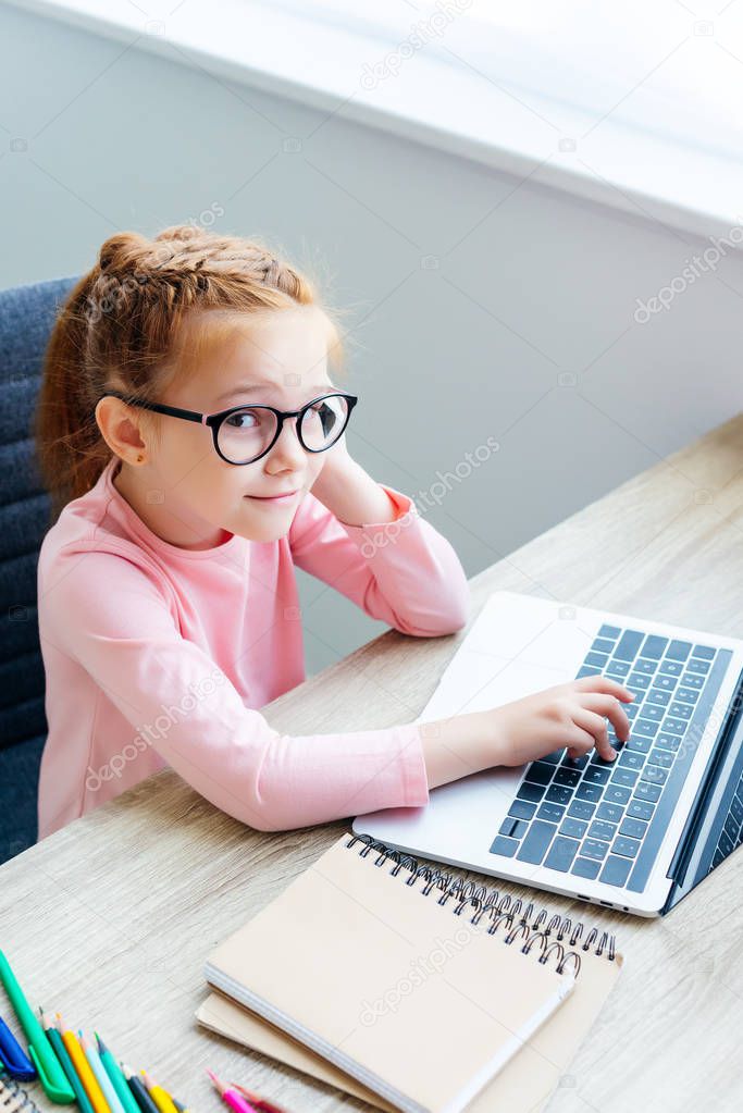 high angle view of smiling schoolchild i eyeglasses using laptop and looking at camera