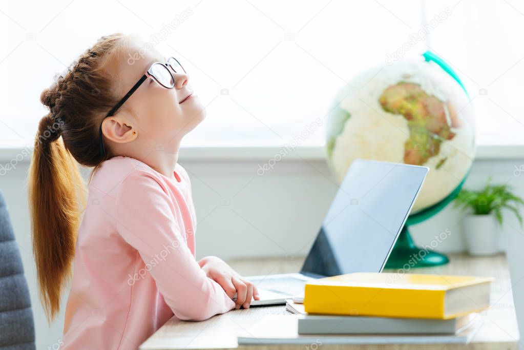 side view of smiling red haired schoolgirl in eyeglasses using laptop at desk