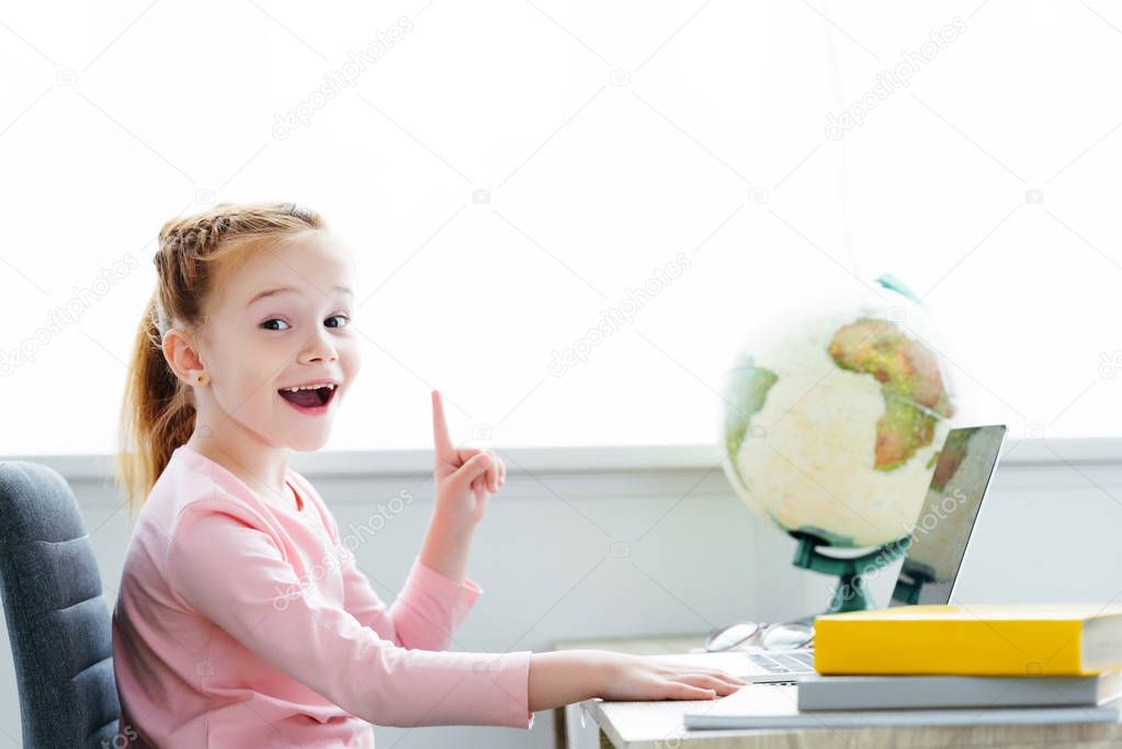 cheerful redhead child pointing up with finger and smiling at camera while studying with books and laptop at home