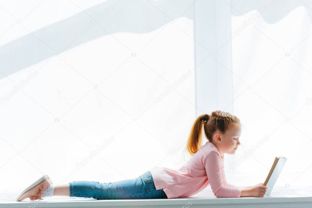 side view of cute little schoolgirl lying on windowsill and reading book 