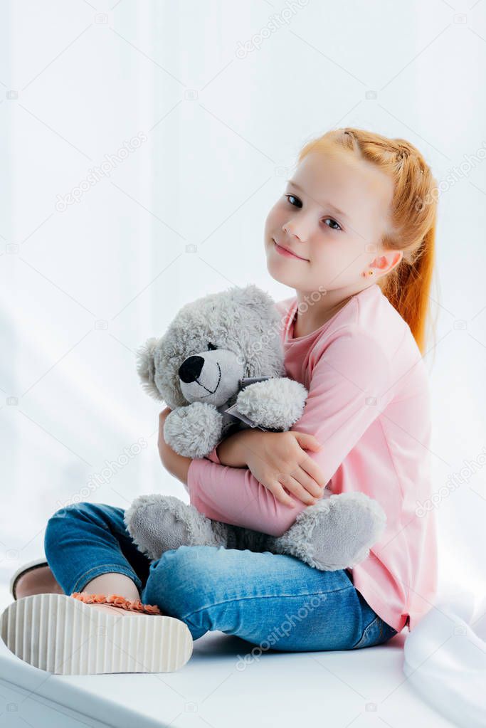 beautiful happy redhead kid hugging teddy bear and smiling at camera