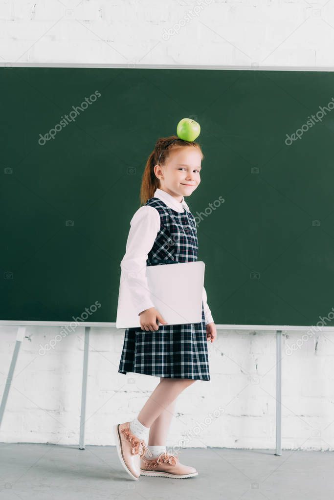 beautiful little schoolgirl with apple on head holding laptop and smiling at camera 
