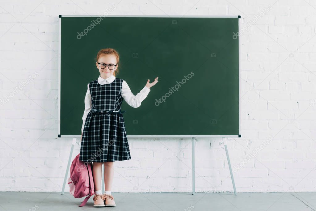 adorable little schoolgirl with backpack showing blank blackboard and smiling at camera 