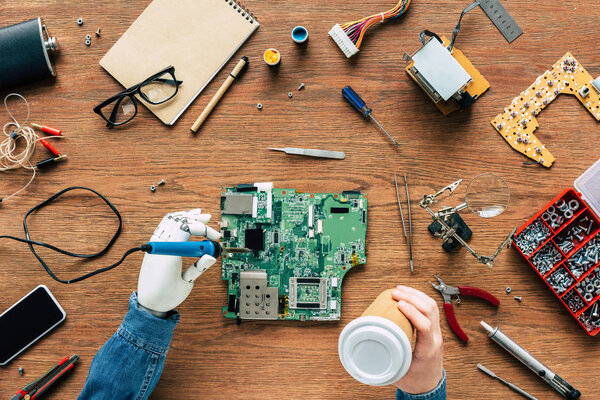 обрезанное изображение инженера-электронщика с протезной паяльной материнской платой и с бумажной чашкой в руках
 