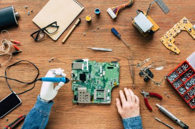 cropped image of electronic engineer with robotic hand fixing motherboard by soldering iron at table surrounded by tools clipart
