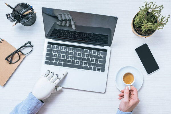 cropped image of businessman with prosthetic arm using laptop and holding coffee cup at table 