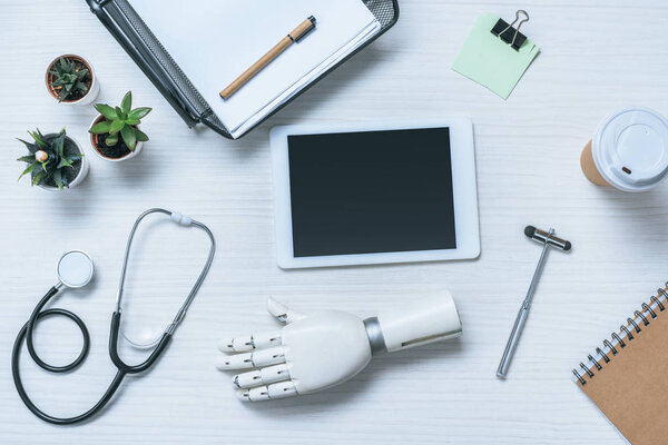 top view of doctor workplace with prosthetic arm, stethoscope, reflex hammer and digital tablet with blank screen on table 