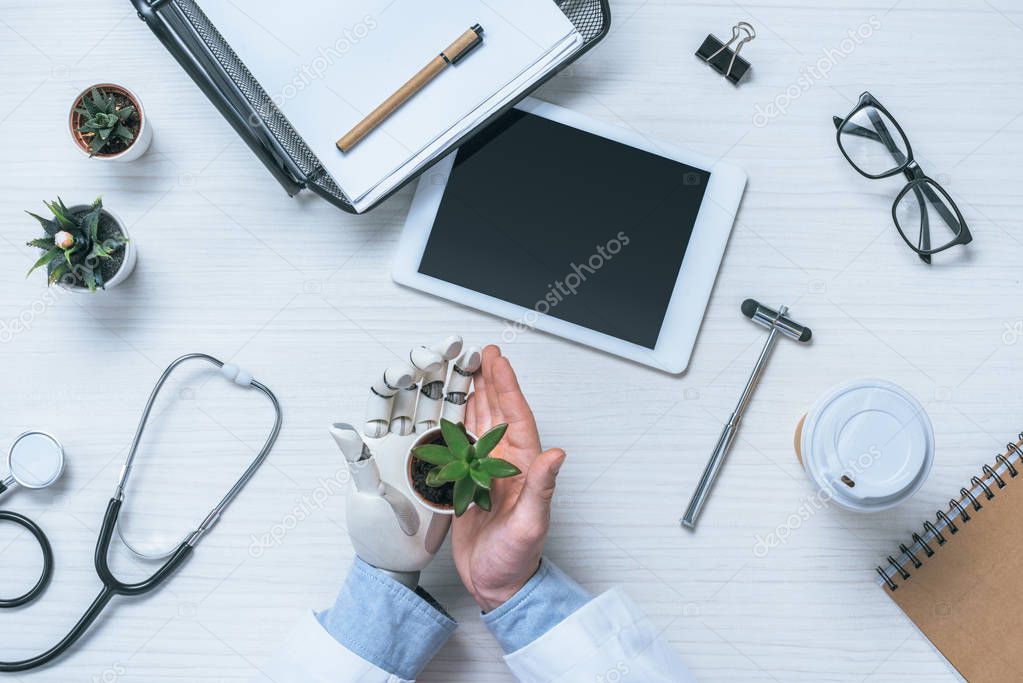 cropped image of male doctor with prosthetic arm holding potted plant at table with medical tools and digital tablet 