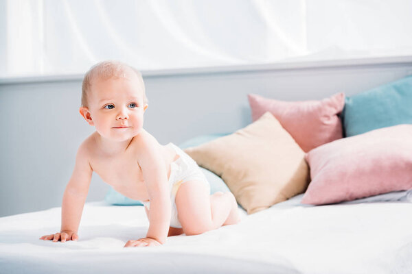 happy little baby crawling in bed with lot of pillows and looking away