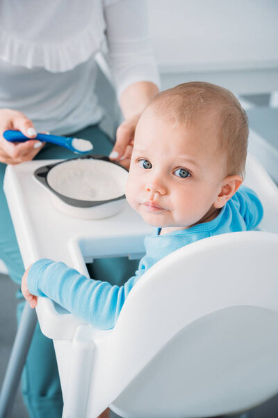 cropped shot of mother feeding adorable child with porridge while he looking at camera