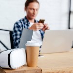 Nahaufnahme von Coffee to go, Virtual-Reality-Headset und Mann mit Smartphone dahinter