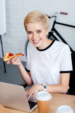 dizüstü bilgisayar kullanarak ve kameraya gülümseyen pizza ile kızın yüksek açılı görünüş