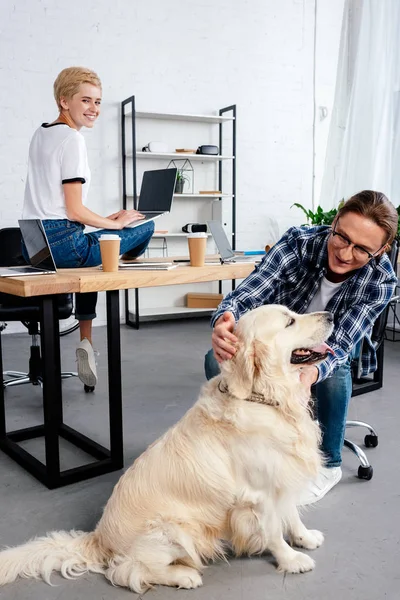 若い女性のラップトップを使用してオフィスで幸せな男なでる犬を見て笑みを浮かべてください  — 無料ストックフォト
