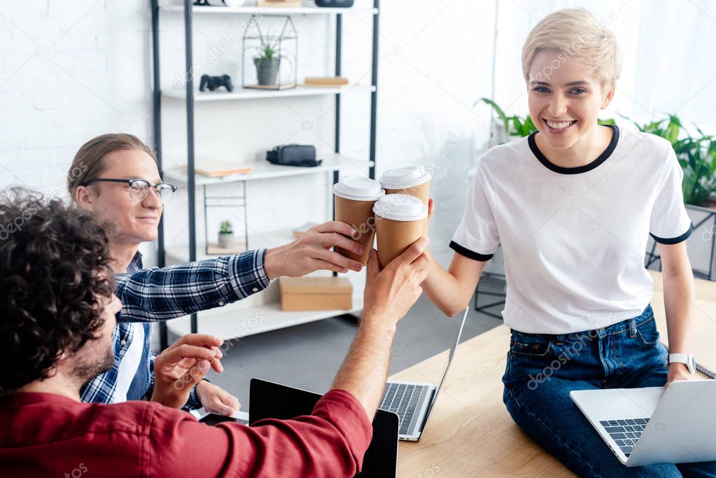 smiling young colleagues clinking paper cups while using laptops in office