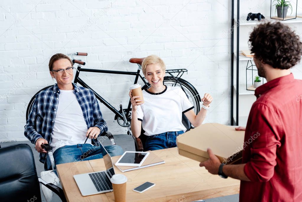 smiling young colleagues looking at man holding pizza boxes in office 