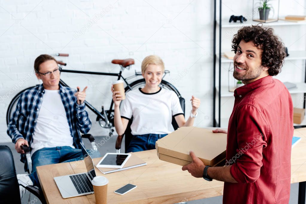 smiling young colleagues looking at happy man holding pizza boxes in office 