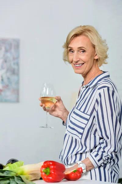 Sonriente Atractiva Mujer Mayor Sosteniendo Vaso Vino Blanco Cocina Mirando — Foto de stock gratuita