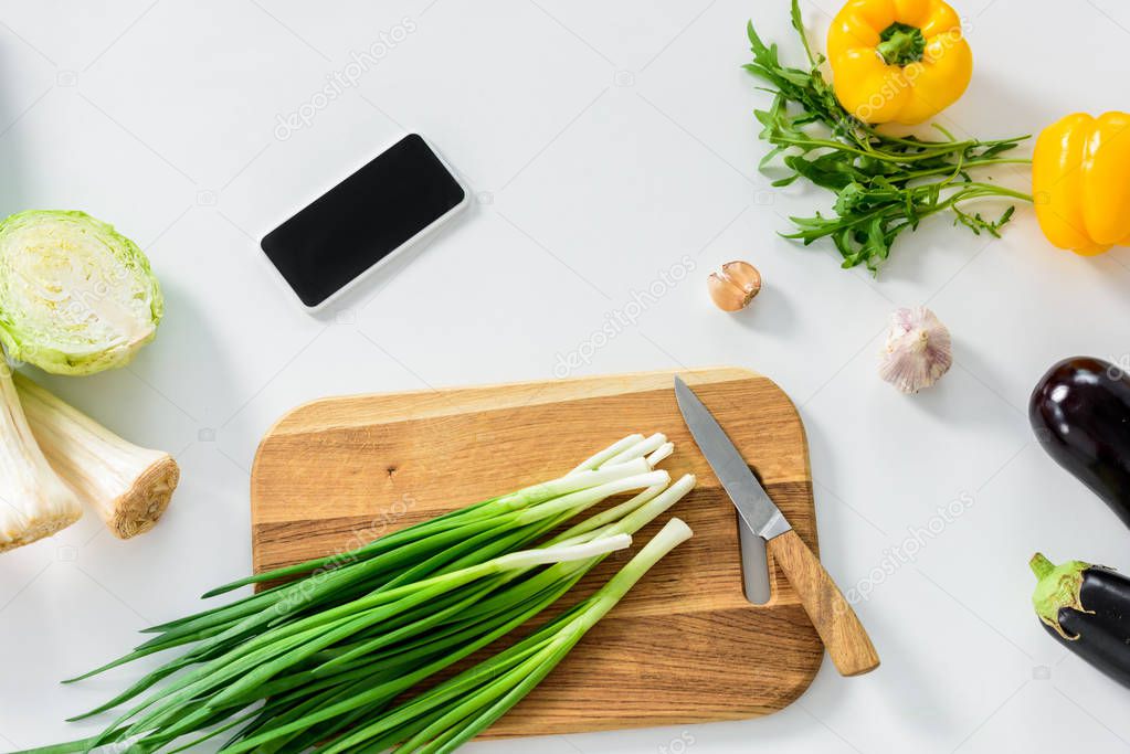 green onion on wooden board, smartphone with blank screen on white tabletop in kitchen