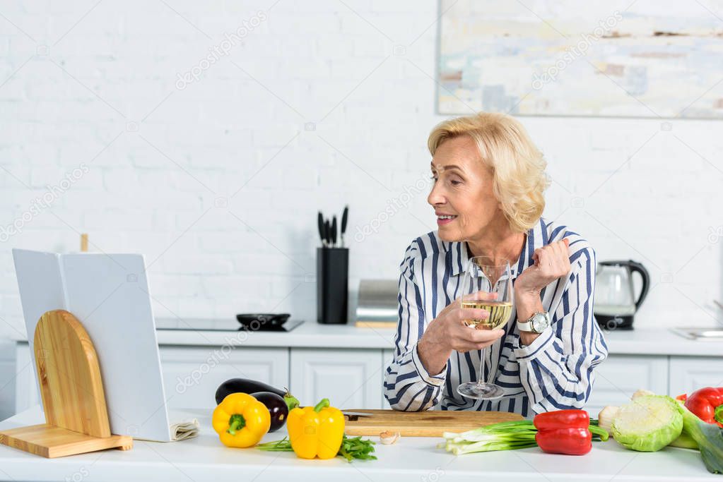 happy attractive grey hair woman holding glass of wine and looking at cookbook in kitchen 