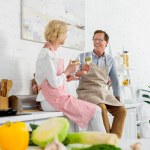 Feliz pareja de ancianos en delantales sosteniendo vasos de vino y sonriéndose en la cocina