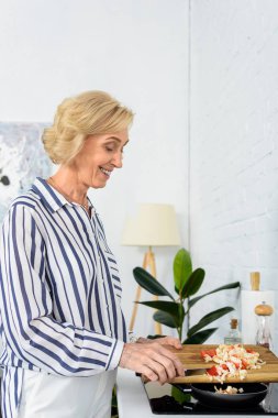 Mutfak tavada sebze koyarak yan görünüm gülümseyen gri saç kadın 