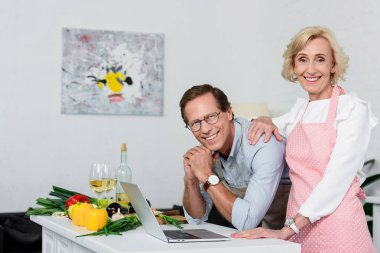 smiling senior couple looking at camera at kitchen clipart