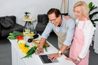 smiling senior couple cooking at kitchen and using laptop clipart