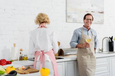 Yaşlı adam şarap tutarak ve karısıyla mutfakta yemek yaparken kameraya gülümseyen önlük