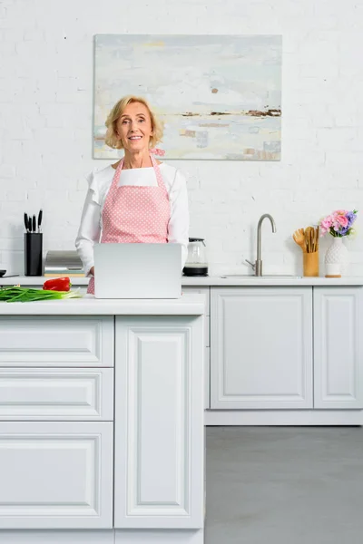 有吸引力的资深妇女使用膝上型电脑烹调在厨房和看照相机 — 免费的图库照片