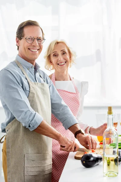 Lächelnder Ehemann Schneidet Gemüse Auf Holzbrett Der Küche Und Blickt — kostenloses Stockfoto