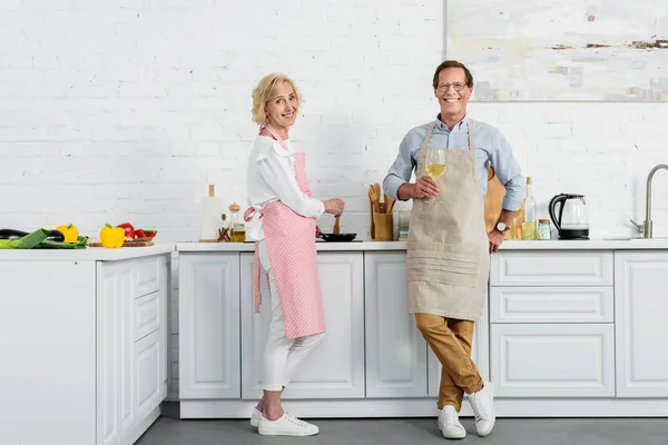 Feliz Pareja Ancianos Delantales Sonriendo Cámara Mientras Bebe Vino Cocina — Foto de Stock