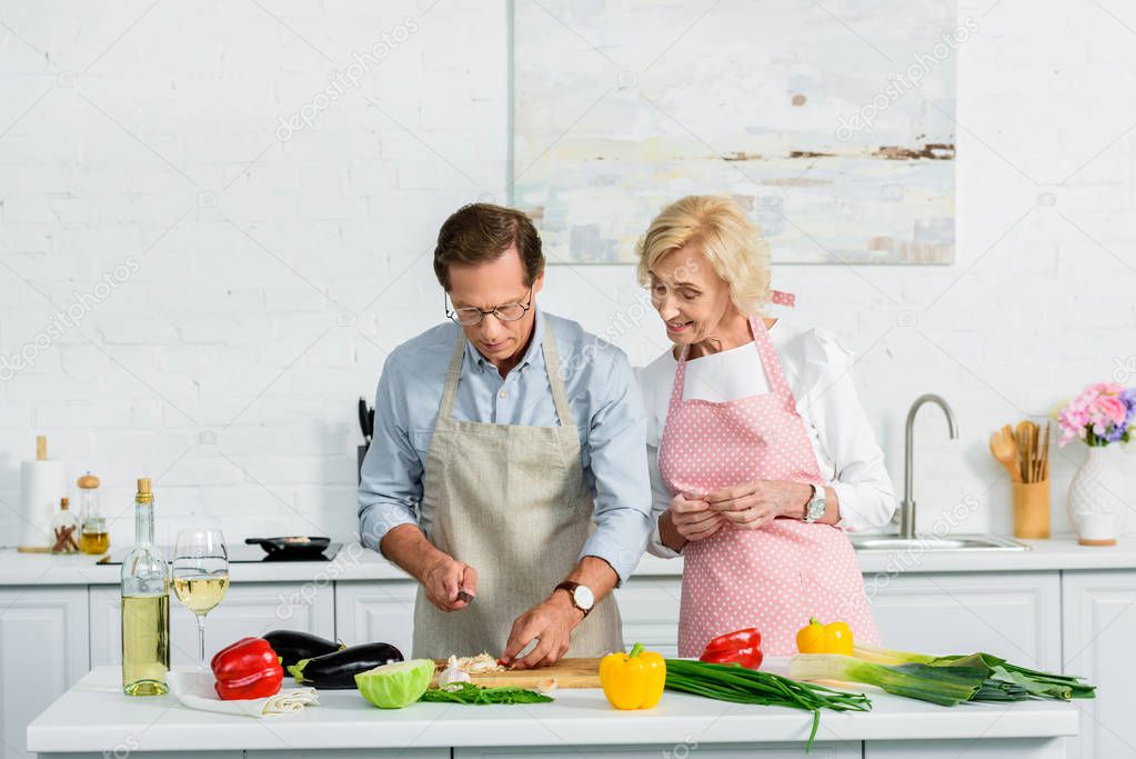 senior wife looking how husband cutting vegetables at kitchen