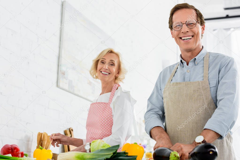 low angle view of happy senior couple cooking together at kitchen and looking at camera