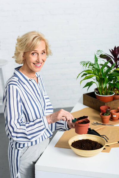 high angle view of happy senior woman holding pot with soil and smiling at camera indoors