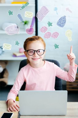 Parmak ile işaret ve eğitim kutsal kişilerin resmi ile evde laptop kullanırken kameraya gülümseyen gözlük içinde çok güzel kırmızı saçlı kız öğrenci