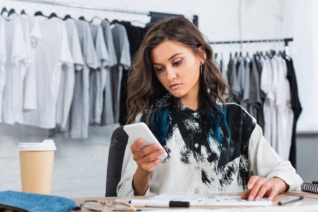 attractive young female designer using smartphone at working table in clothing design studio