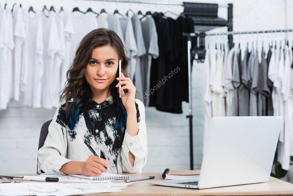 young female fashion designer talking on smartphone at working table and looking at camera in clothing design studio