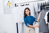 smiling female designer taking grey t-shirt from hanger in clothing design studio