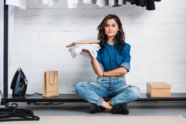 smiling female designer sitting with stack of white t-shirts and paper package in clothing design studio