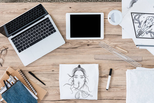 top view of t-shirt with print, laptop and digital tablet with blank screens at working table 
