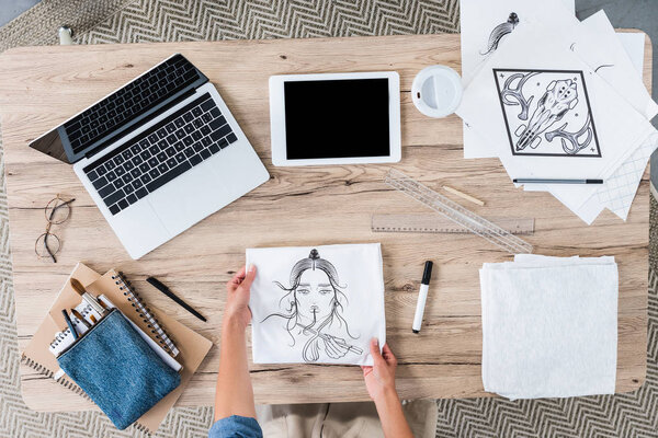 cropped image of female designer putting white t-shirt with print on table with digital devices an paintings 