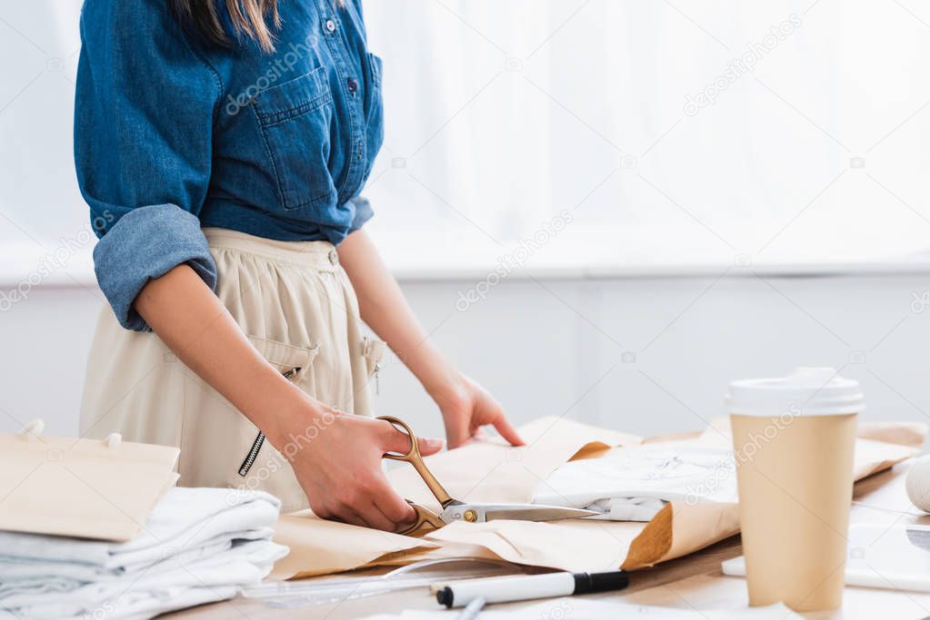 cropped image of female fashion designer wrapping t-shirt with print in paper package at table with coffee cup