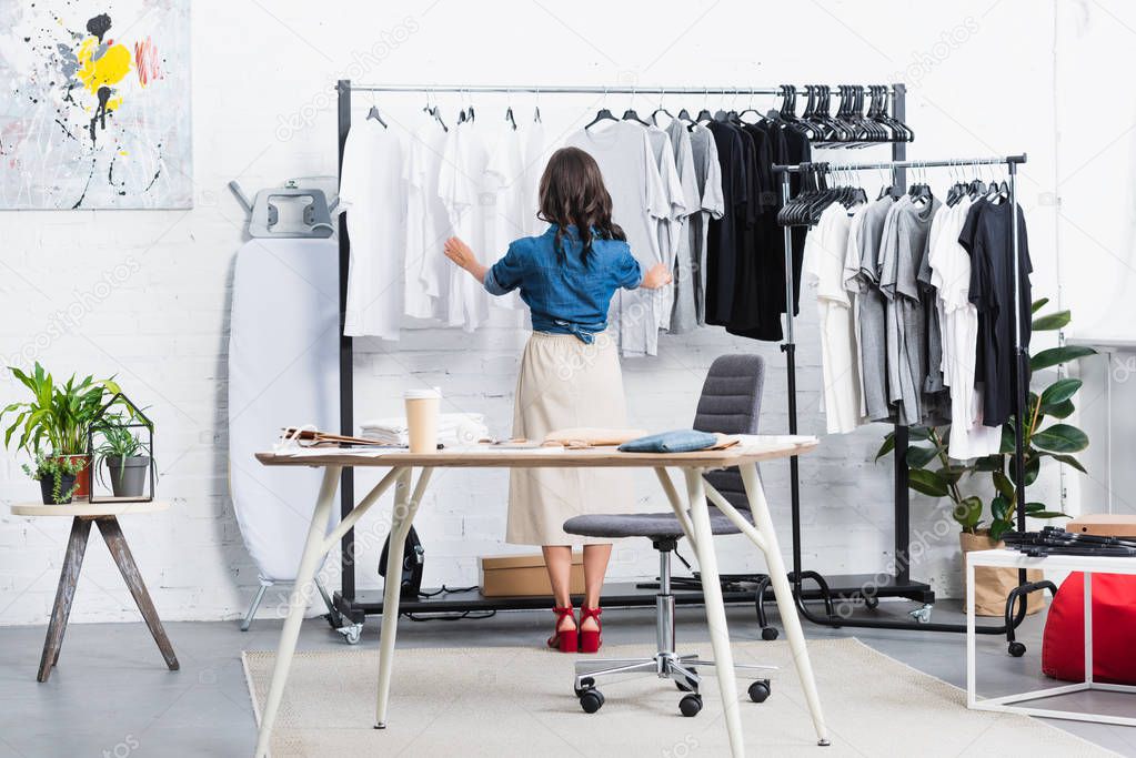 rear view of female designer taking black t-shirt from hanger in clothing design studio