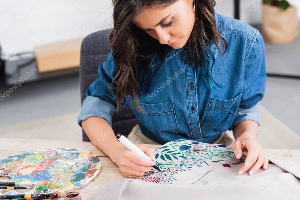 selective focus of female fashion designer painting on jacket at working table in clothing design studio