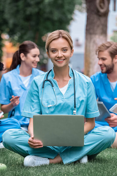 улыбающийся студент-медик учится с ноутбуком на зеленой траве
