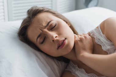 attractive young woman suffering from pain in neck while lying in bed clipart