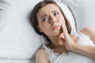 attractive young woman suffering from toothache and looking away while lying in bed  clipart